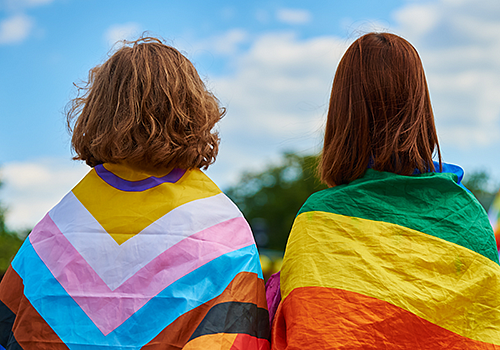 Two students with their backs turned, drapped in rainbow flags