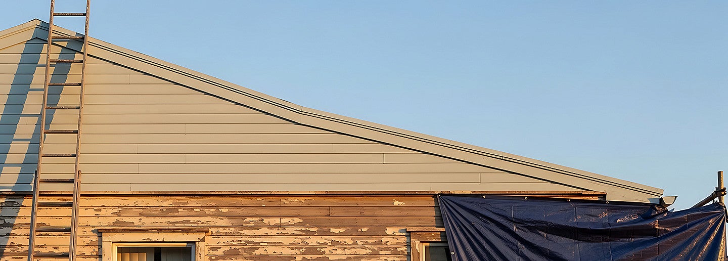 ixer upper house exterior showing old wood siding with peeling paint and a ladder against the new roof during a residential home renovation project